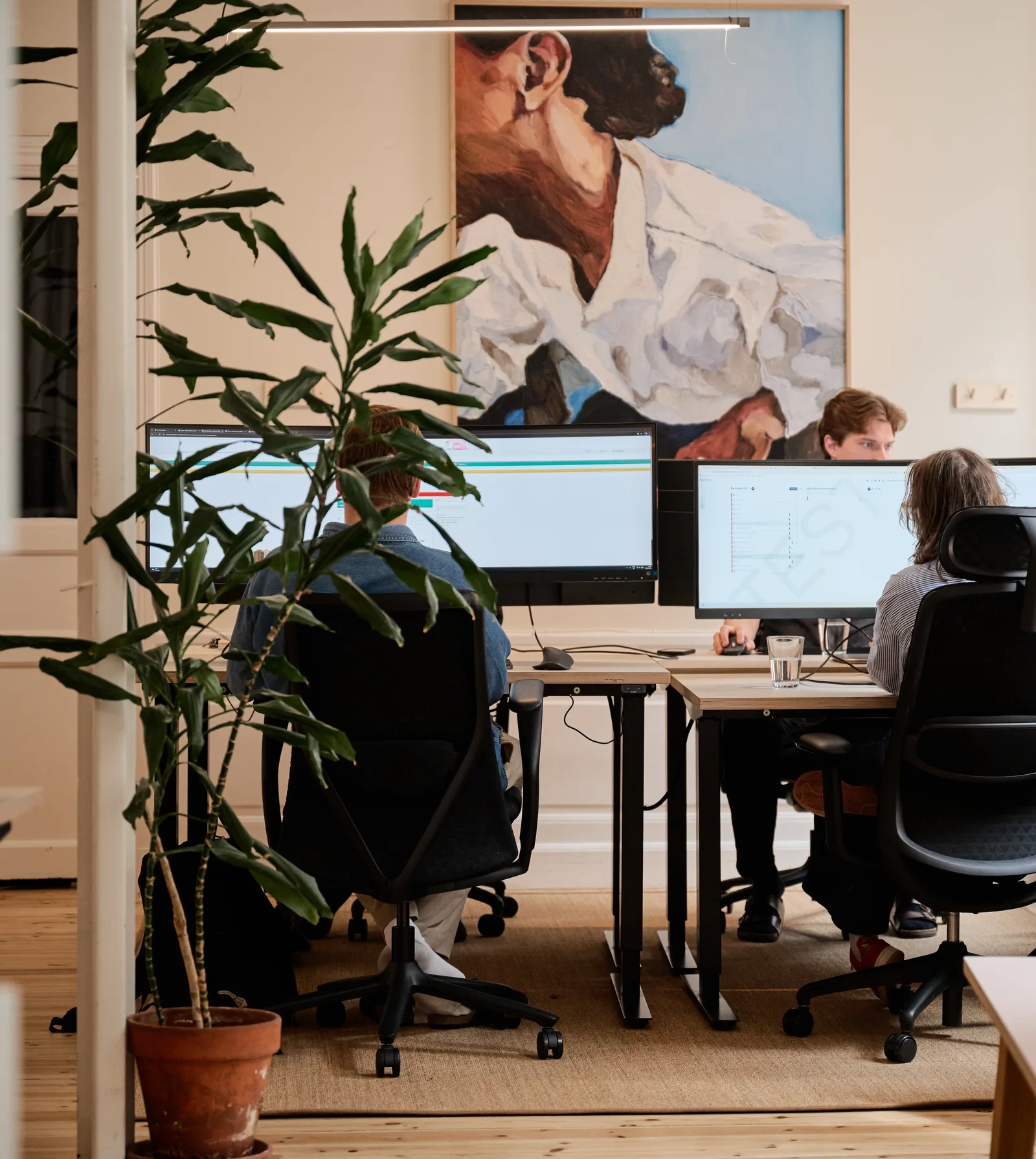 Vy genom en dörröppning mot kontorets arbetsplatser där tre konsulter sitter vid sina skärmar under en stor målning